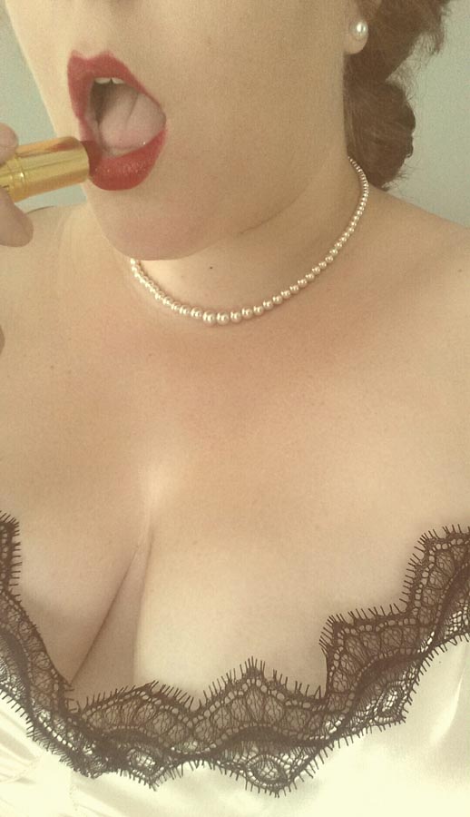 A woman in a negligee and pearls applying red lipstick.