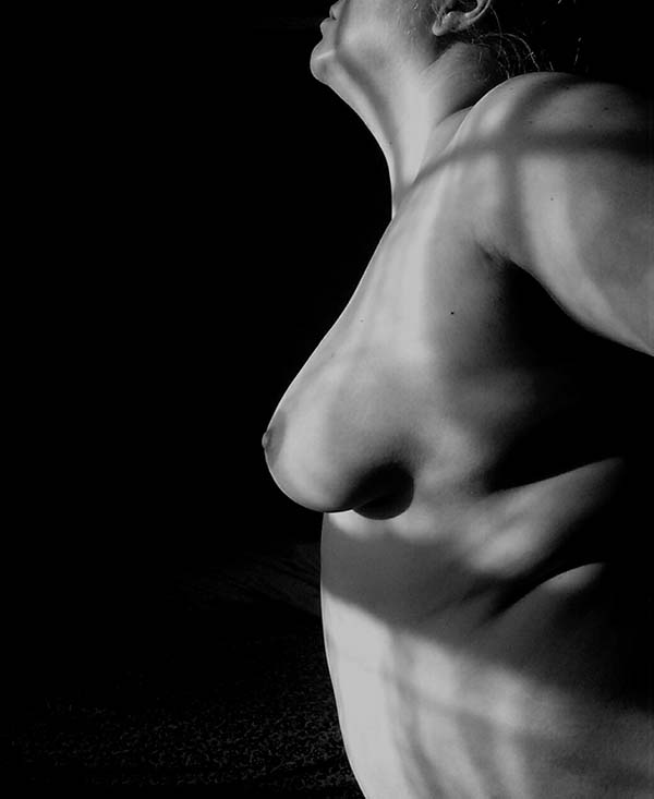 The profile of a nude woman, light from a window shining across her breasts, her head and shoulders back. Black and white.