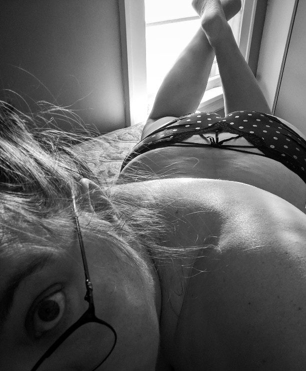 black and white photo of woman in polka dot panties laying on bed, looking at camera, wearing glasses